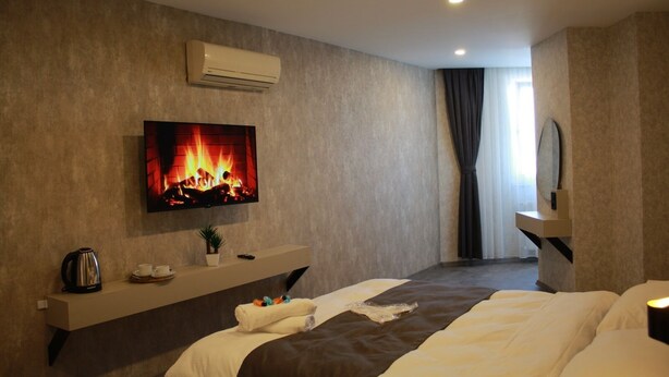 A modern hotel room with a flat-screen TV displaying a fireplace, a bedside table with a kettle and cups, a bed with a brown headboard, and a large window with curtains.