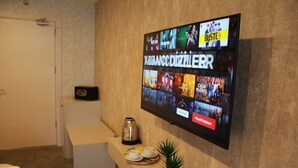 A hotel reception area with a flat-screen TV displaying various shows and movies. There is a coffee maker, a small potted plant, and a tray with cups and saucers on a counter.