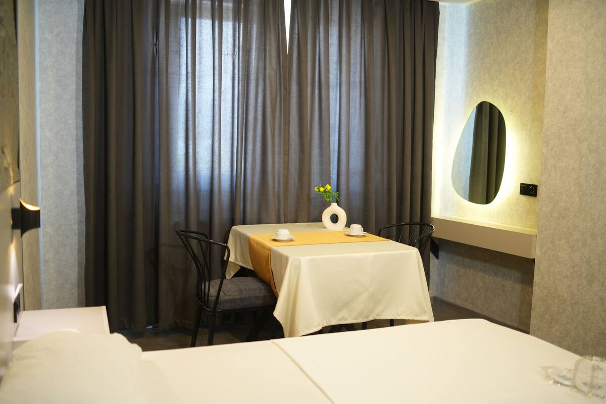 Ett hotellrum med en snyggt bäddad säng, ett matbord med ett vitt bordsduks, en vas med blommor och en spegel på väggen.