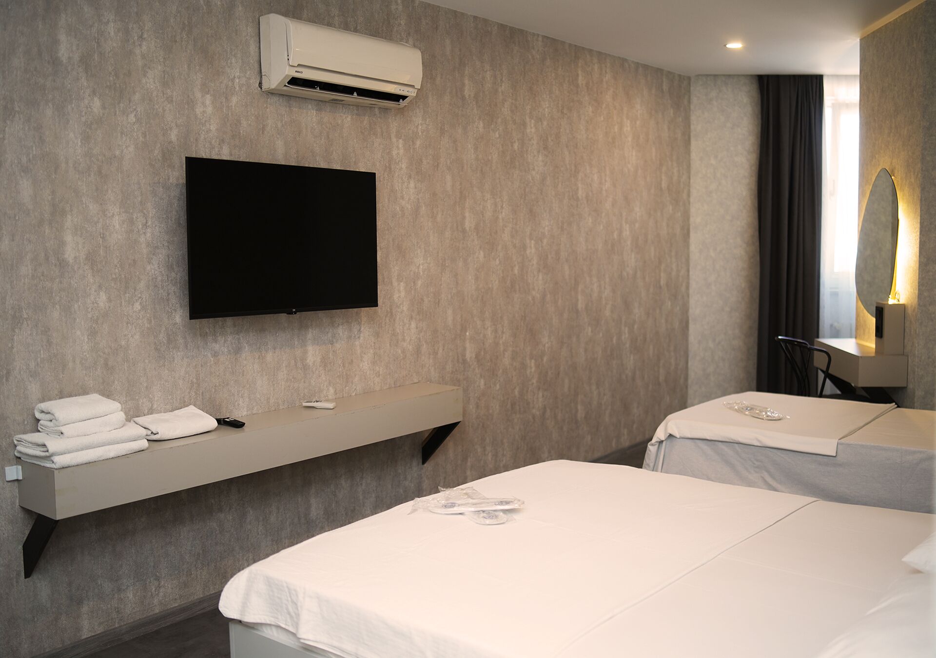 Habitación de hotel con televisión de pantalla plana, aire acondicionado integrado, tocador con espejo y dos camas con ropa blanca.