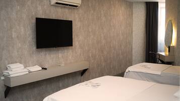 Habitación de hotel con televisión de pantalla plana, aire acondicionado integrado, tocador con espejo y dos camas con ropa blanca.