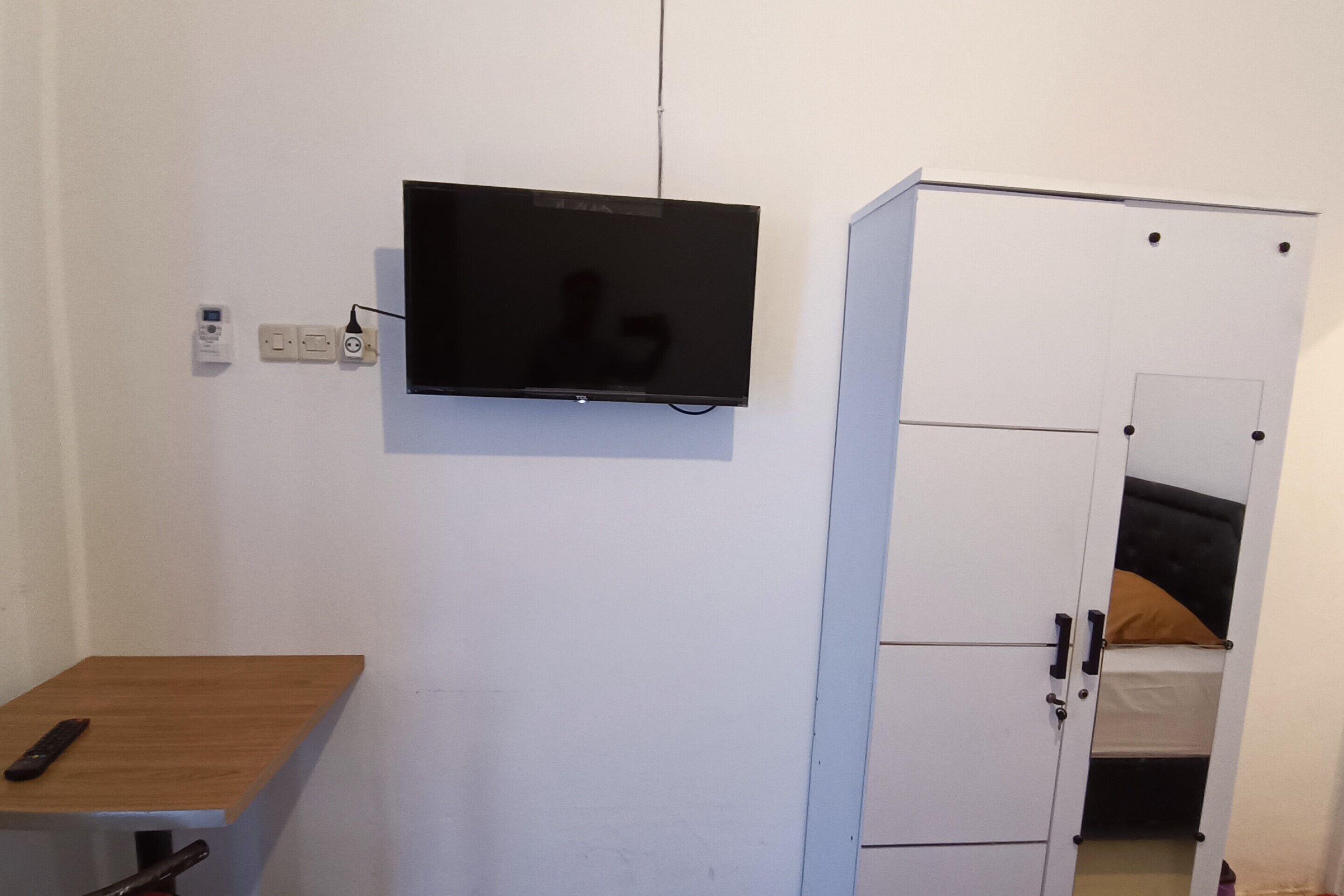 A room with a TV mounted on the wall, a wooden table, and a white wardrobe with a bed in the background.