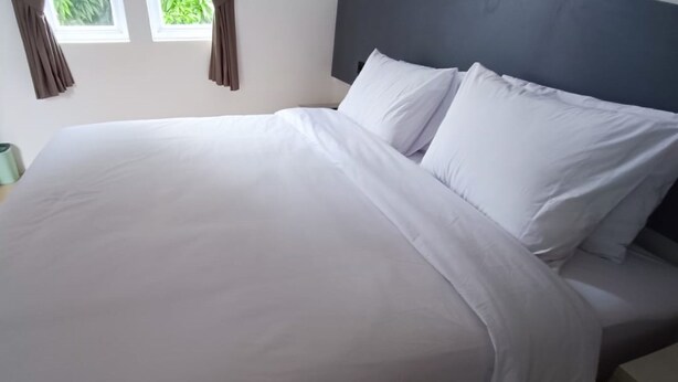 A neatly made bed with white linens and pillows, a grey headboard, and a window with curtains. - Hotel O Bumi Tenang Homestay (Salatiga)