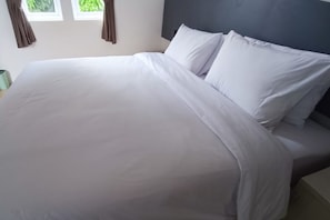 Een netjes opgemaakt bed met witte lakens en kussens, een grijs hoofdbord en een raam met gordijnen.