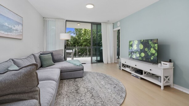 A modern living room with a grey sectional sofa, a round rug, a flat-screen TV on a white stand, and a balcony with a view of trees and a building. - Blissful Bribie Beach Haven (WOORIM)