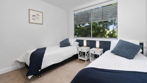 A bedroom with two single beds, a small bedside table with a lamp, a framed picture on the wall, and a window with blinds. - Blissful Bribie Beach Haven (WOORIM)