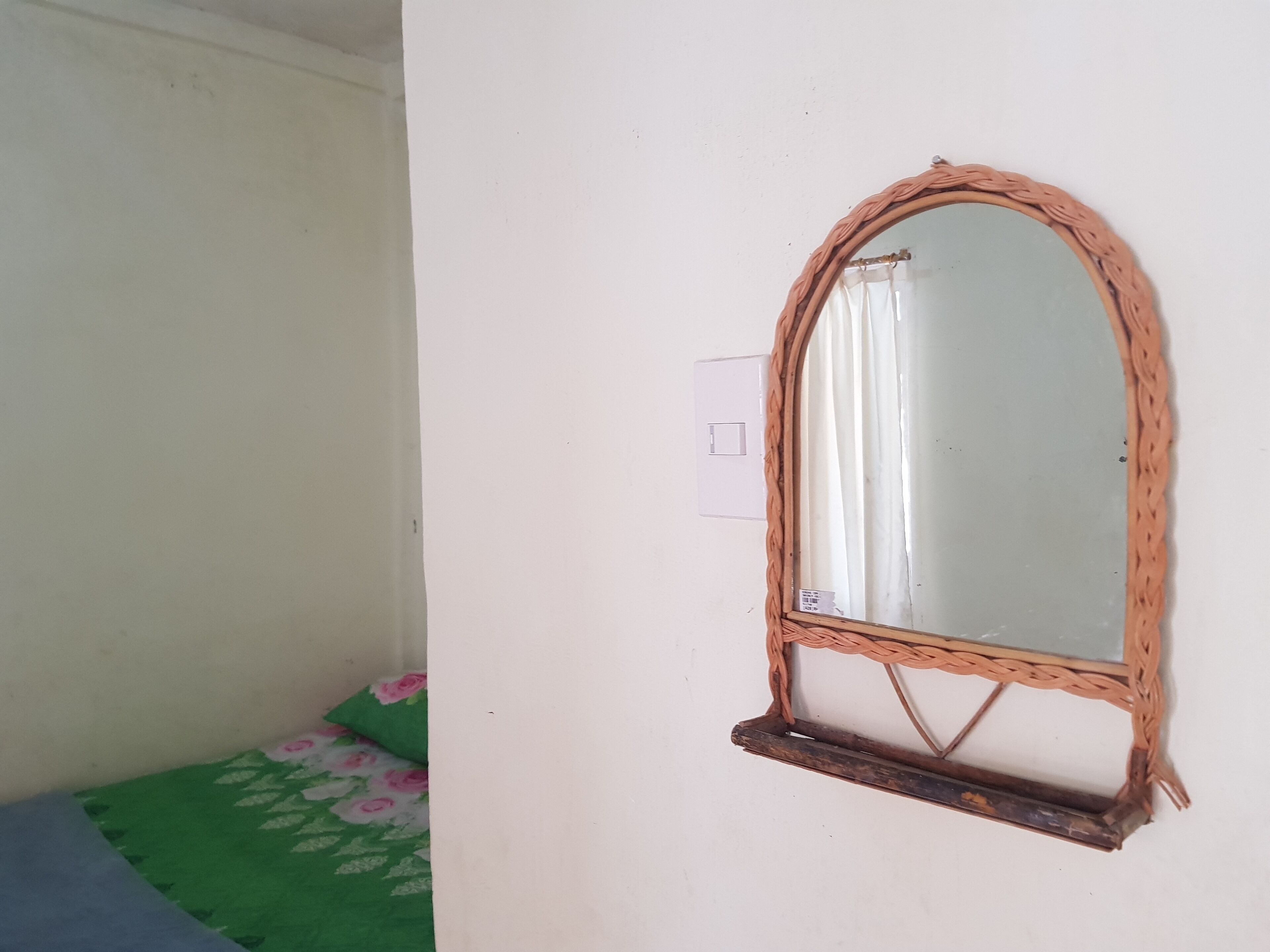 Habitación de esquina con un espejo de marco tejido en la pared, un enchufe y una colcha de cama estampada con motivos florales verdes.