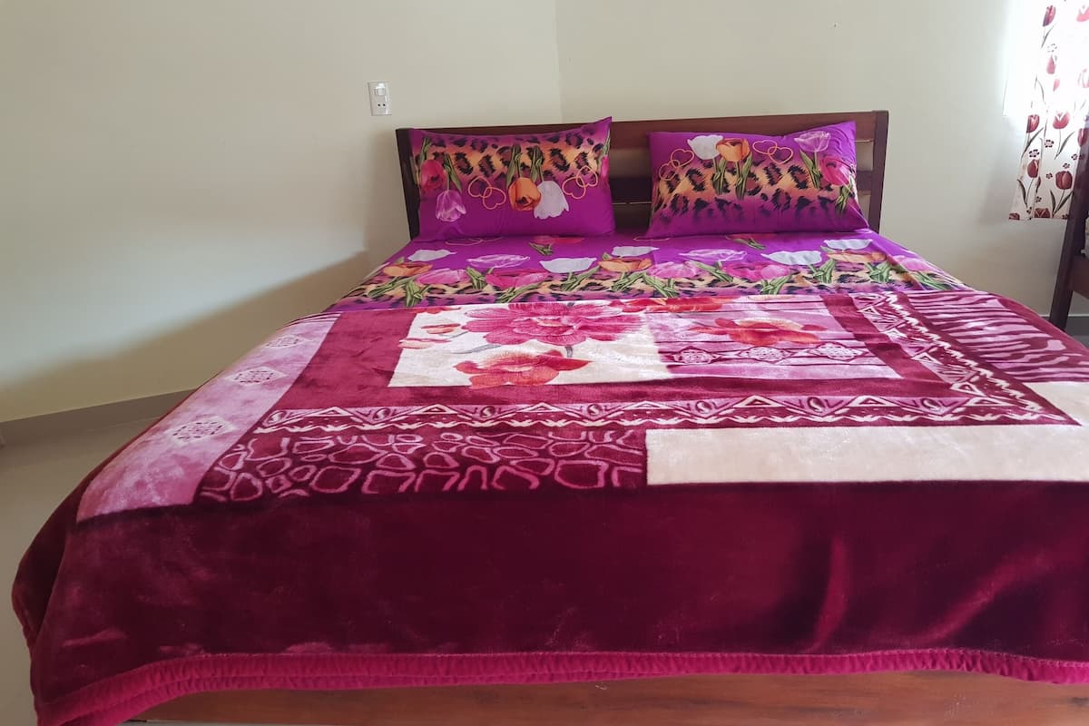 A bed with a floral-patterned bedspread and two pillows, a wooden headboard, and a plain wall with a power outlet.