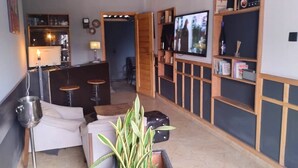 A modern living room with a sofa, a coffee table, and a TV. There are shelves with books and decorative items, and a small bar area with stools. - The Crib-Artist Residence (Port Harcourt)