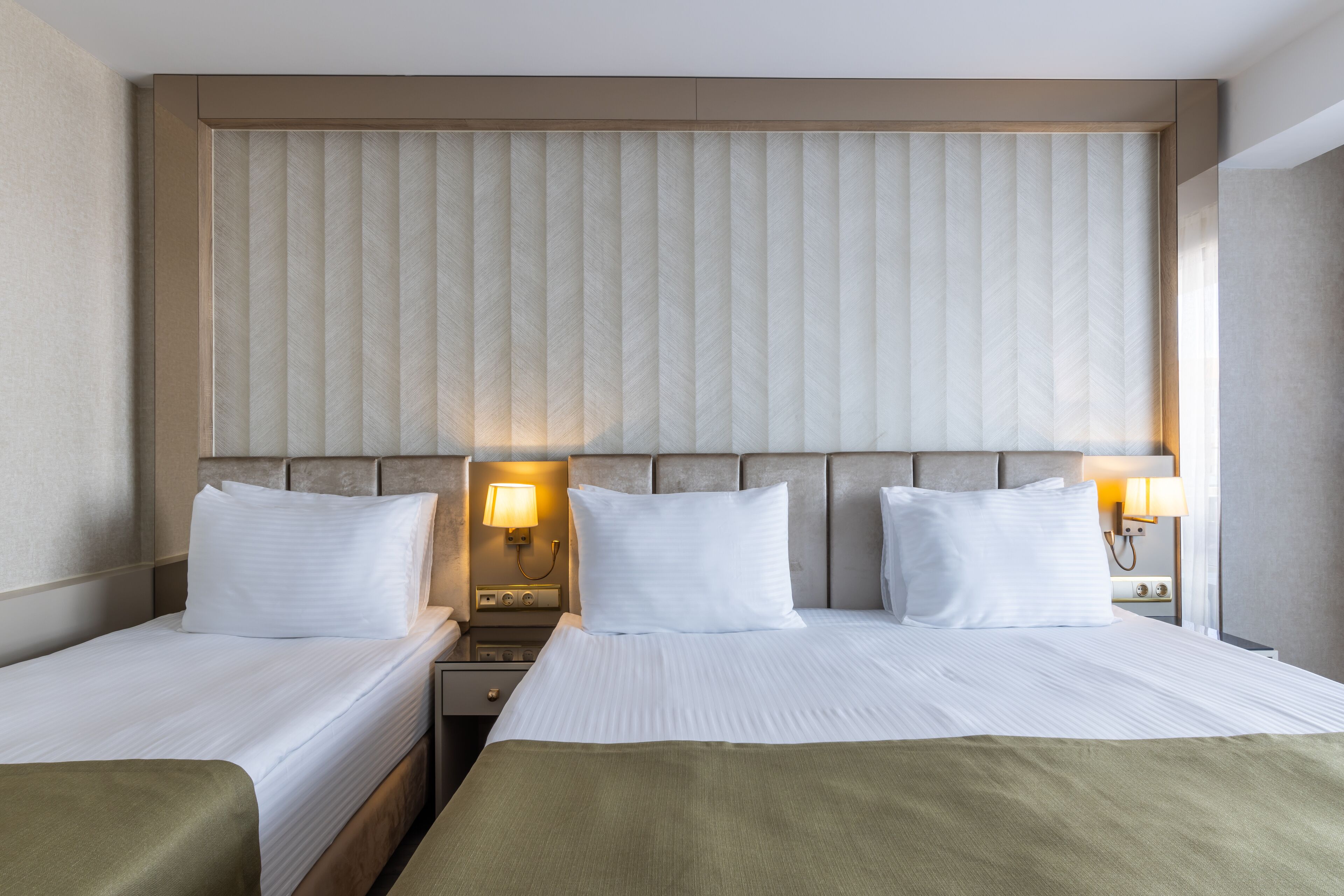 Ein Hotelzimmer mit zwei Betten, einem Kopfteil mit vertikalen Jalousien, Nachttischlampen und einer neutralen Farbgestaltung.