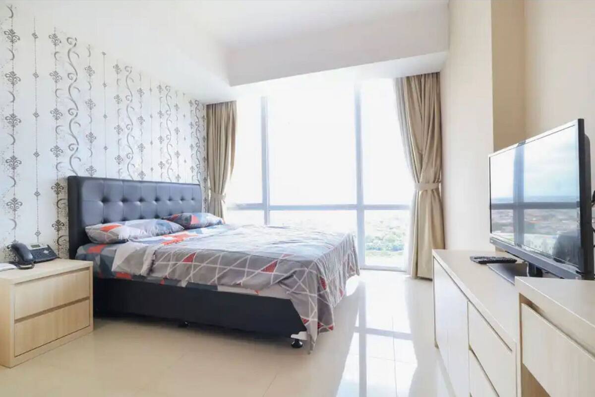 A modern bedroom with a large bed, a bedside table with a phone, a TV on a stand, and a view of the city through large windows.