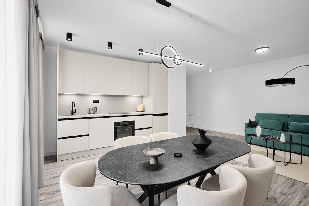 Una cocina moderna con armarios blancos, una mesa de comedor con sillas, un sofá verde y una lámpara colgante negra.
