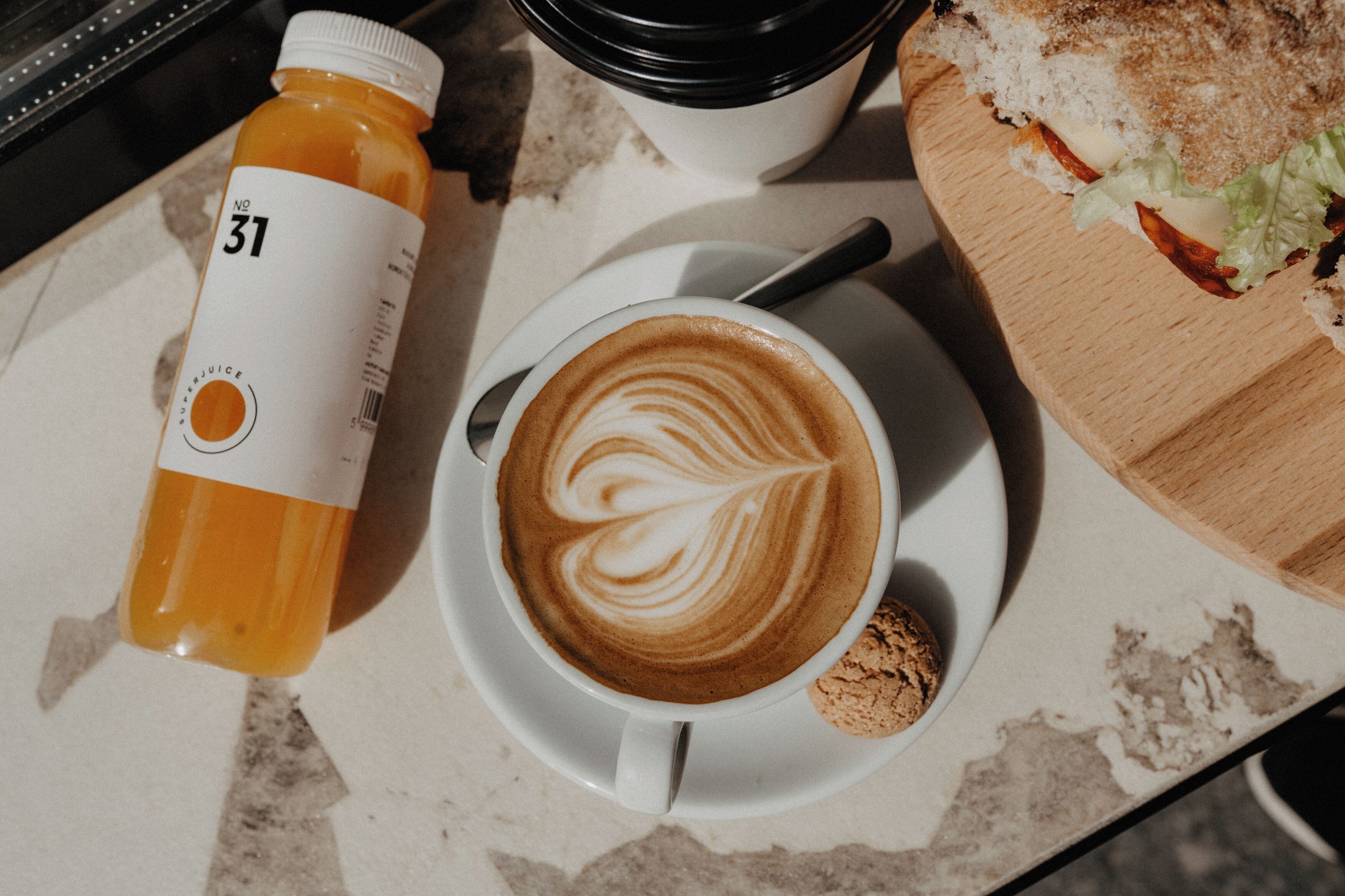 A cup of coffee with latte art on a saucer, a bottle of orange juice, and a sandwich on a wooden board.
