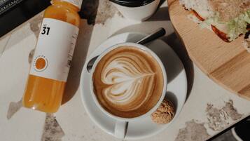 A cup of coffee with latte art on a saucer, a bottle of orange juice, and a sandwich on a wooden board.