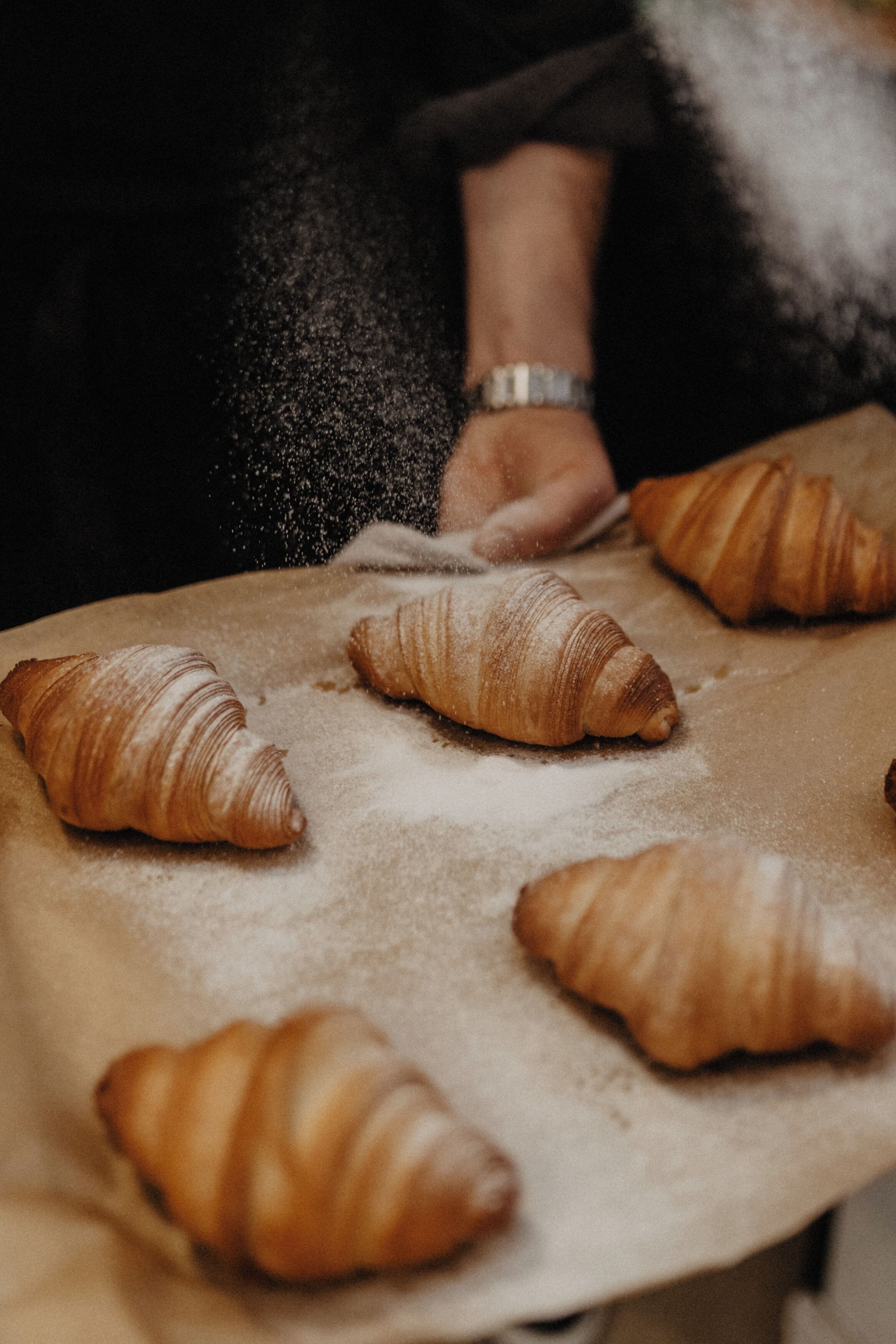 Una persona espolvoreando harina sobre croissants en una bandeja para hornear.