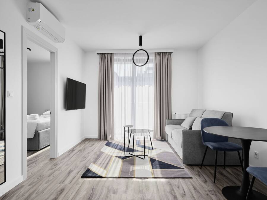 A modern hotel room with a sofa, a round dining table, a small stool, a TV mounted on the wall, and a large window with curtains.
