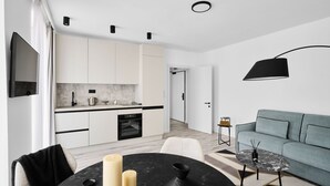 Cocina moderna con armarios blancos, encimera negra y horno empotrado. Hay un comedor con mesa y sillas negras, y un sofá gris en la sala de estar.