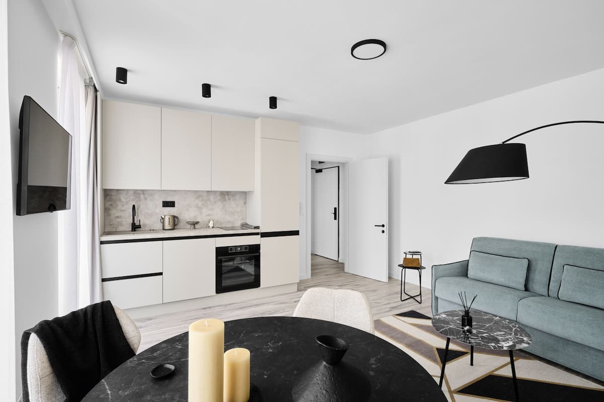 Cocina moderna con armarios blancos, encimera negra y horno empotrado. Hay un comedor con mesa y sillas negras, y un sofá gris en la sala de estar.
