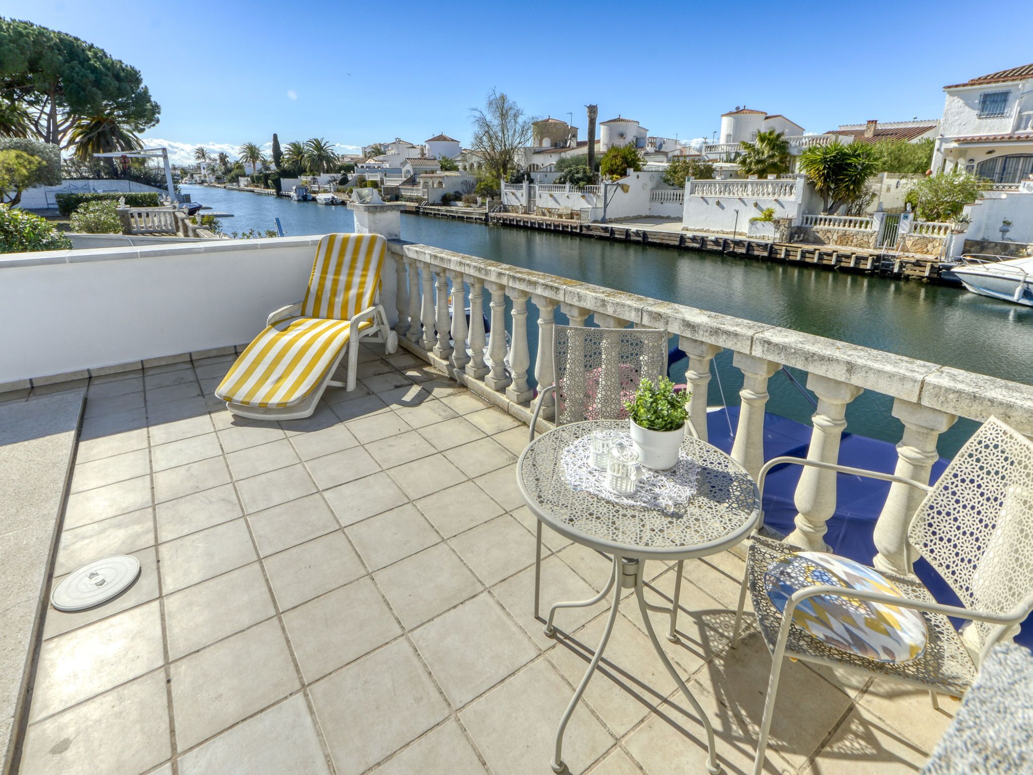 Une terrasse avec un sol carrelé, deux fauteuils de jardin à rayures, une petite table ronde agrémentée d’un pot de fleurs, et une balustrade donnant sur une étendue d’eau où l’on aperçoit des bateaux et des bâtiments au loin.