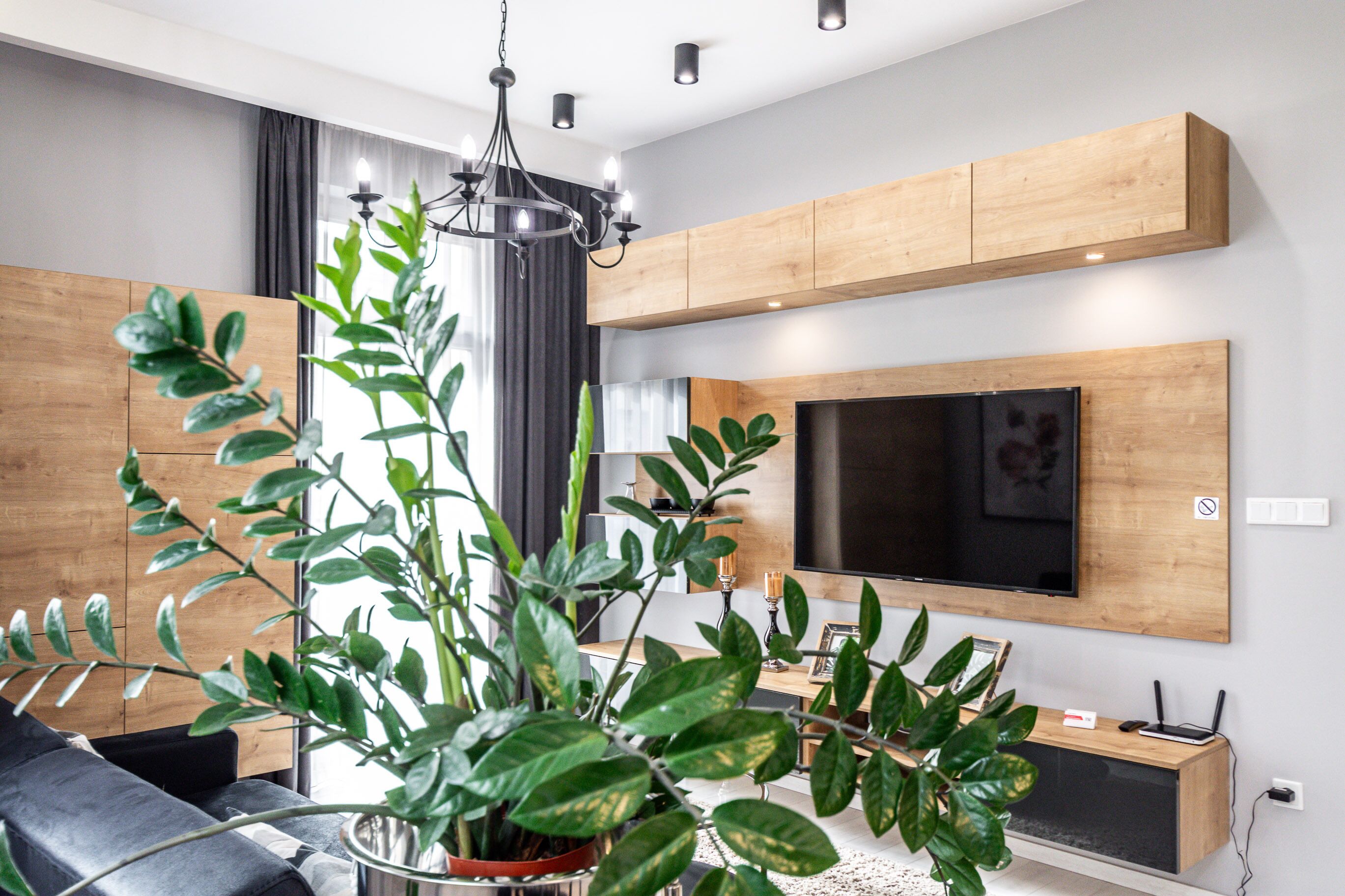 Ein modernes Wohnzimmer mit einem großen Flachbildfernseher an einer Holzwand, einer Kristallleuchtre und einem Blumentopf.
