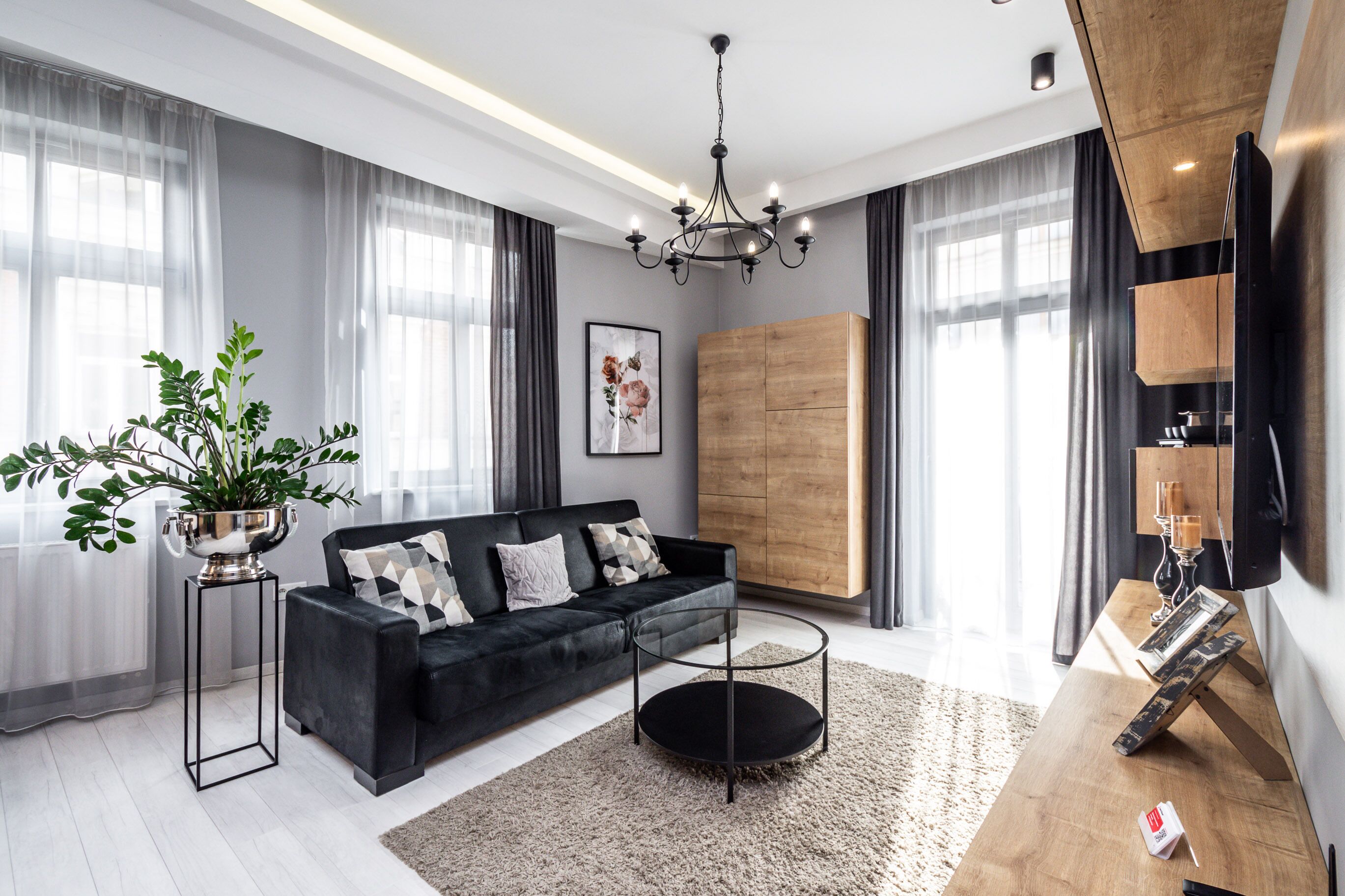 Una sala de estar moderna con un sofá negro, un mueble de madera, una araña y un ventanal con cortinas translúcidas.