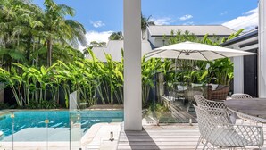 A poolside area with a wooden deck, white wicker chairs, and a large umbrella. - Byron Bay Accom - Sunset on Massinger - 60 Massinger Street - Pets on Request (Byron Bay)