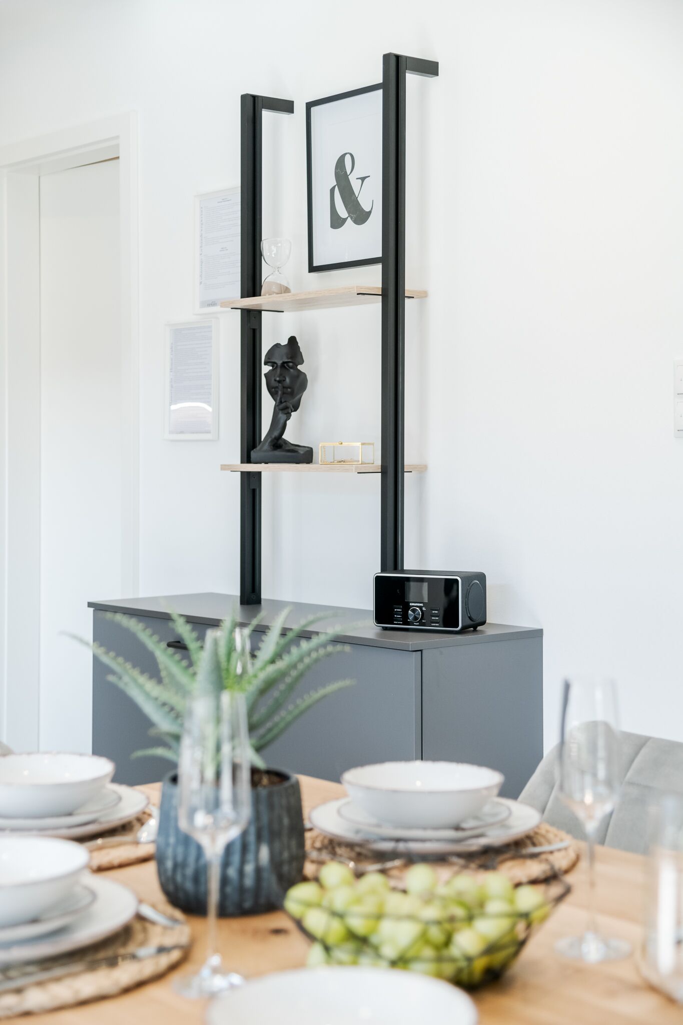 Un espace repas moderne avec une table au design minimaliste, une étagère décorative en noir et blanc et une petite radio posée sur une commode grise.