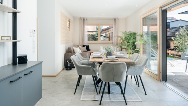 Uma zona de refeições moderna, com uma mesa posta para quatro pessoas, um sofá e uma porta de vidro deslizante que dá para um espaço ao ar livre.