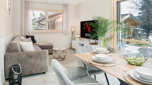 Uma sala de estar moderna com um sofá, mesa de jantar posta com pratos e copos, uma televisão e uma janela grande com cortinas.