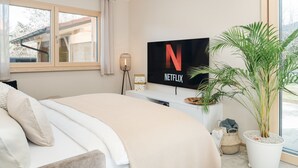 Um quarto moderno com uma cama grande, uma TV na parede exibindo o logotipo da Netflix, uma planta em vaso e uma janela com cortinas.