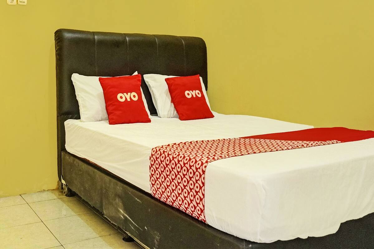 Een bed met een zwart hoofdeinde, witte beddengoed en een rode en witte gestreepte plaid.