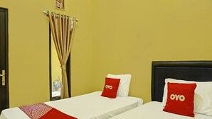Habitación de hotel con dos camas individuales, cada una con un colchón blanco y una almohada roja con la palabra 'OYO'. Las paredes son amarillas y hay una ventana con cortinas.