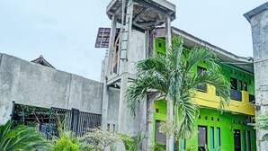 Un edificio de varios pisos con fachada verde y amarilla, rodeado de abundante vegetación y una estructura de concreto.