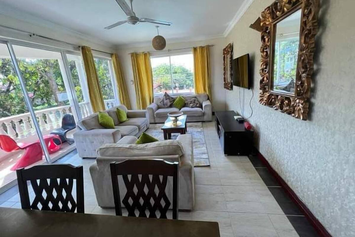 A spacious living room with a large sofa set, a dining table, and chairs. There is a ceiling fan, a TV mounted on the wall, and a balcony with a view of greenery.