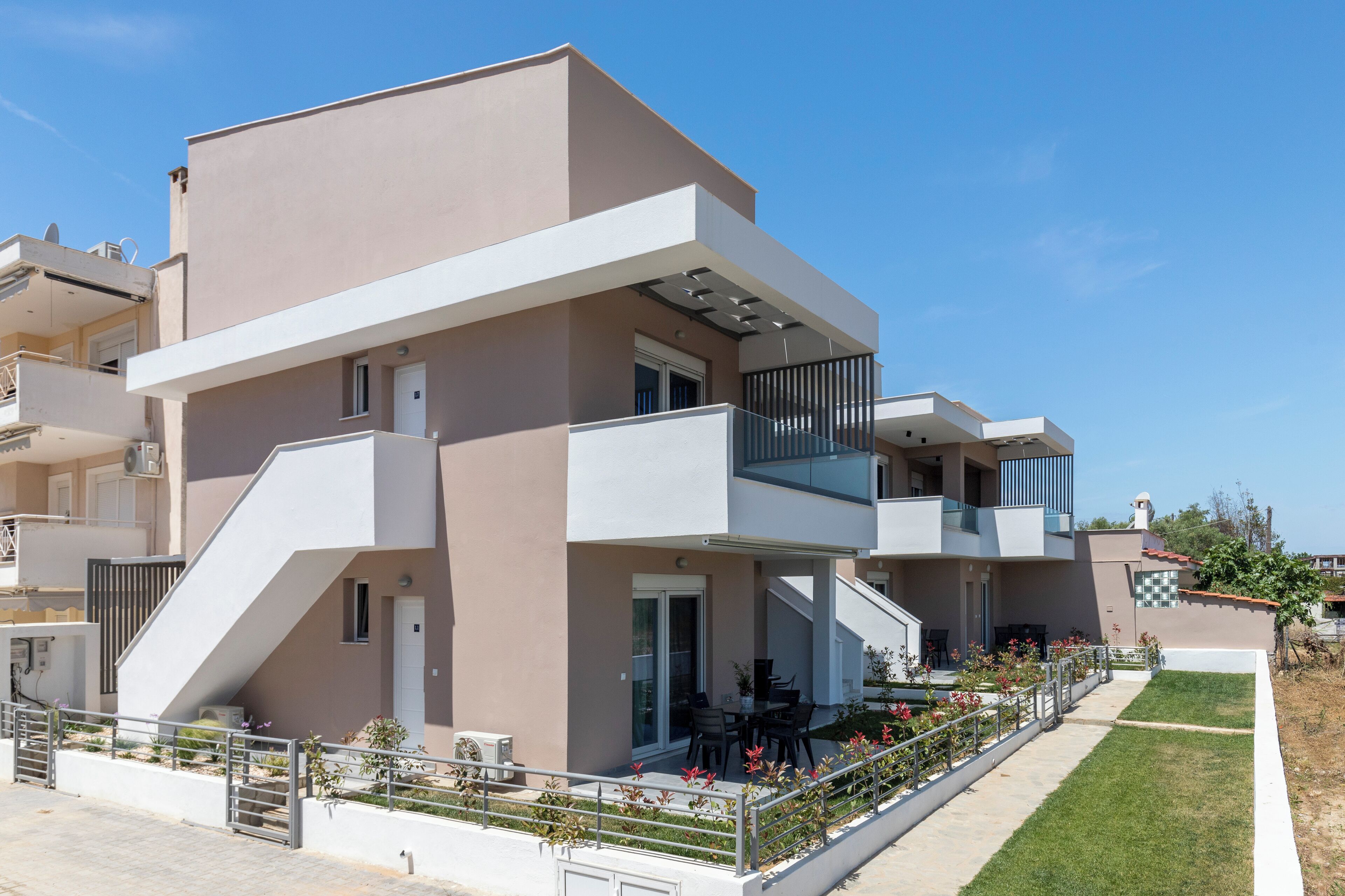 A modern two-story residential building with a flat roof, large windows, and a balcony with a metal railing.