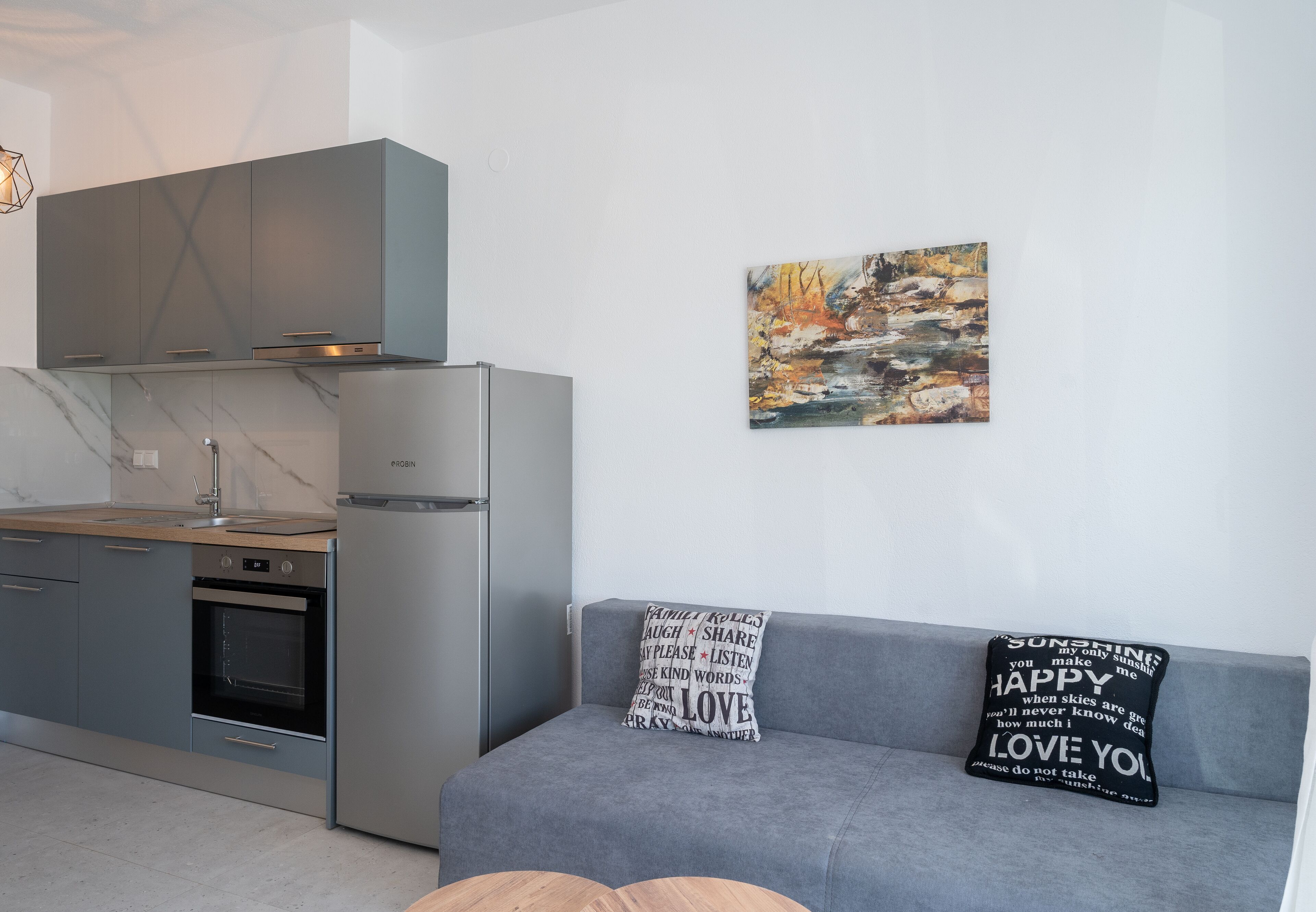 Et moderne køkken med grå skabe, en indbygget ovn og et rustfrit stål køleskab. Der er en grå sofa med to dekorative puder og et maleri af et landskab på væggen.