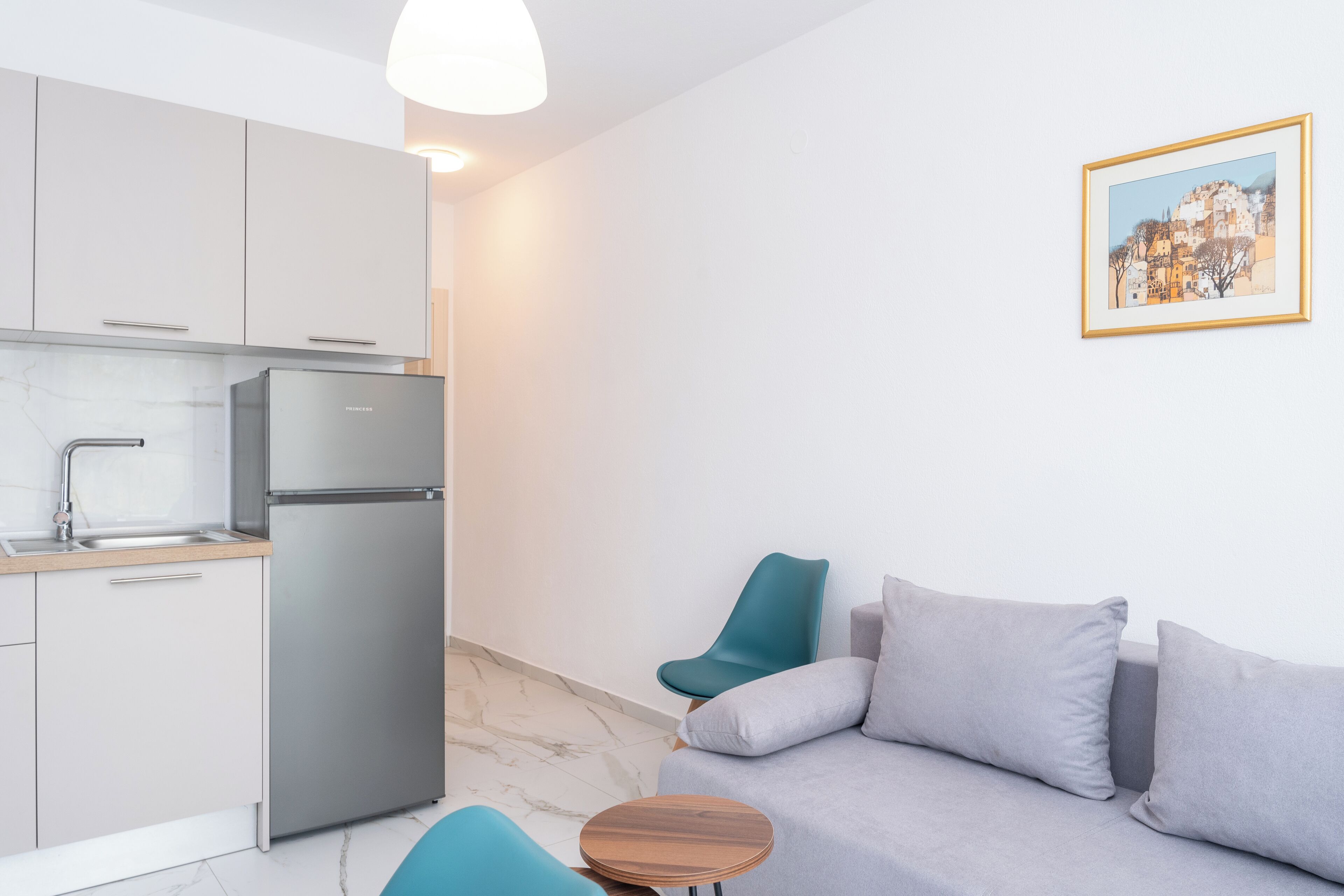 A modern kitchen with a refrigerator, a sink, and a wooden table. There is a gray sofa with teal chairs and a framed picture on the wall.