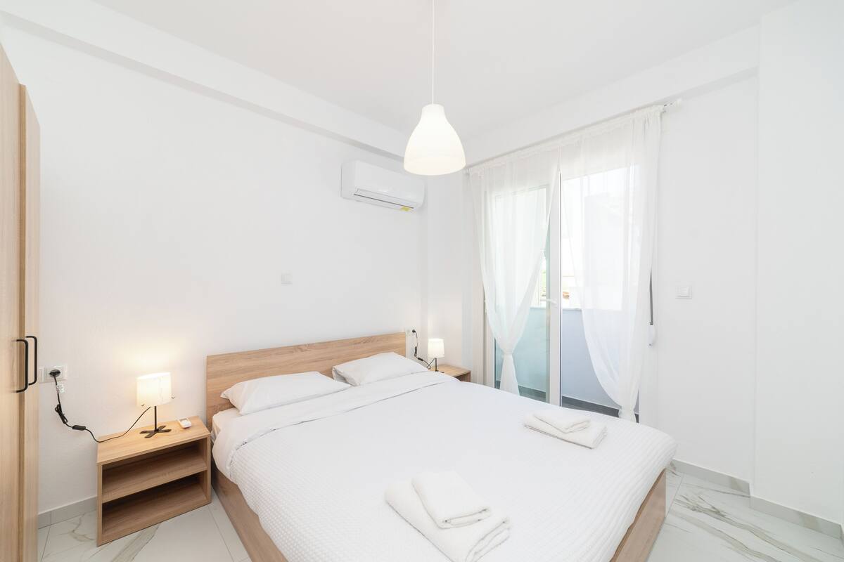 Una habitación de hotel moderna con una cama grande, mesitas de noche con lámparas, un equipo de aire acondicionado y vistas al exterior a través de cortinas translúcidas.