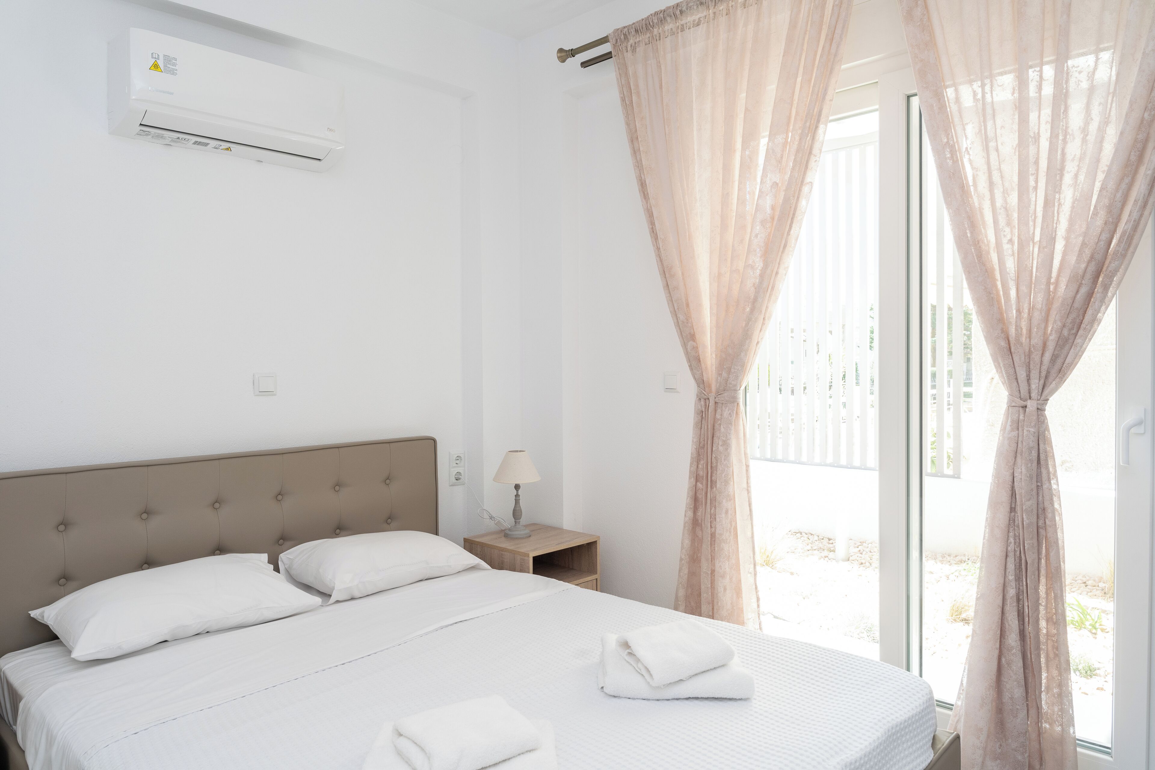 A hotel room with a bed, a bedside table with a lamp, and a window with sheer curtains.