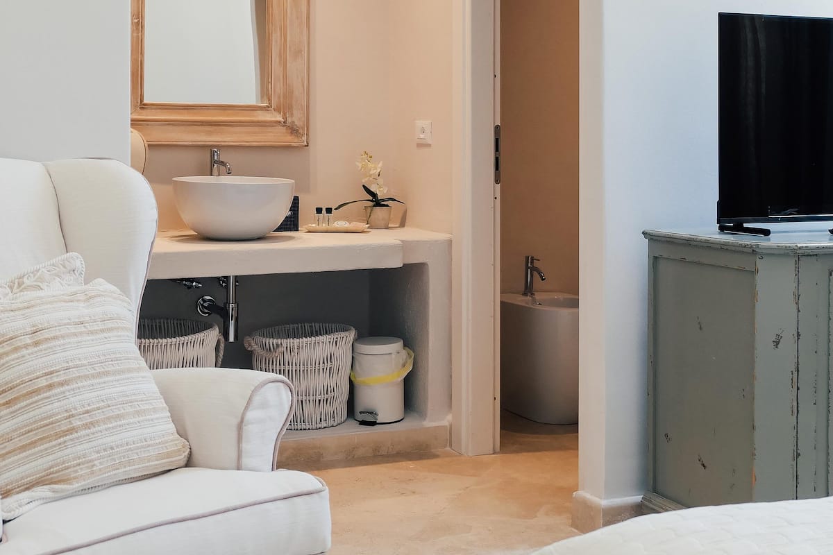 Ein Badezimmer mit einem weißen Waschbecken, Spiegel und WC, ein weißes Sofa sowie ein Fernseher auf einem Schrank.