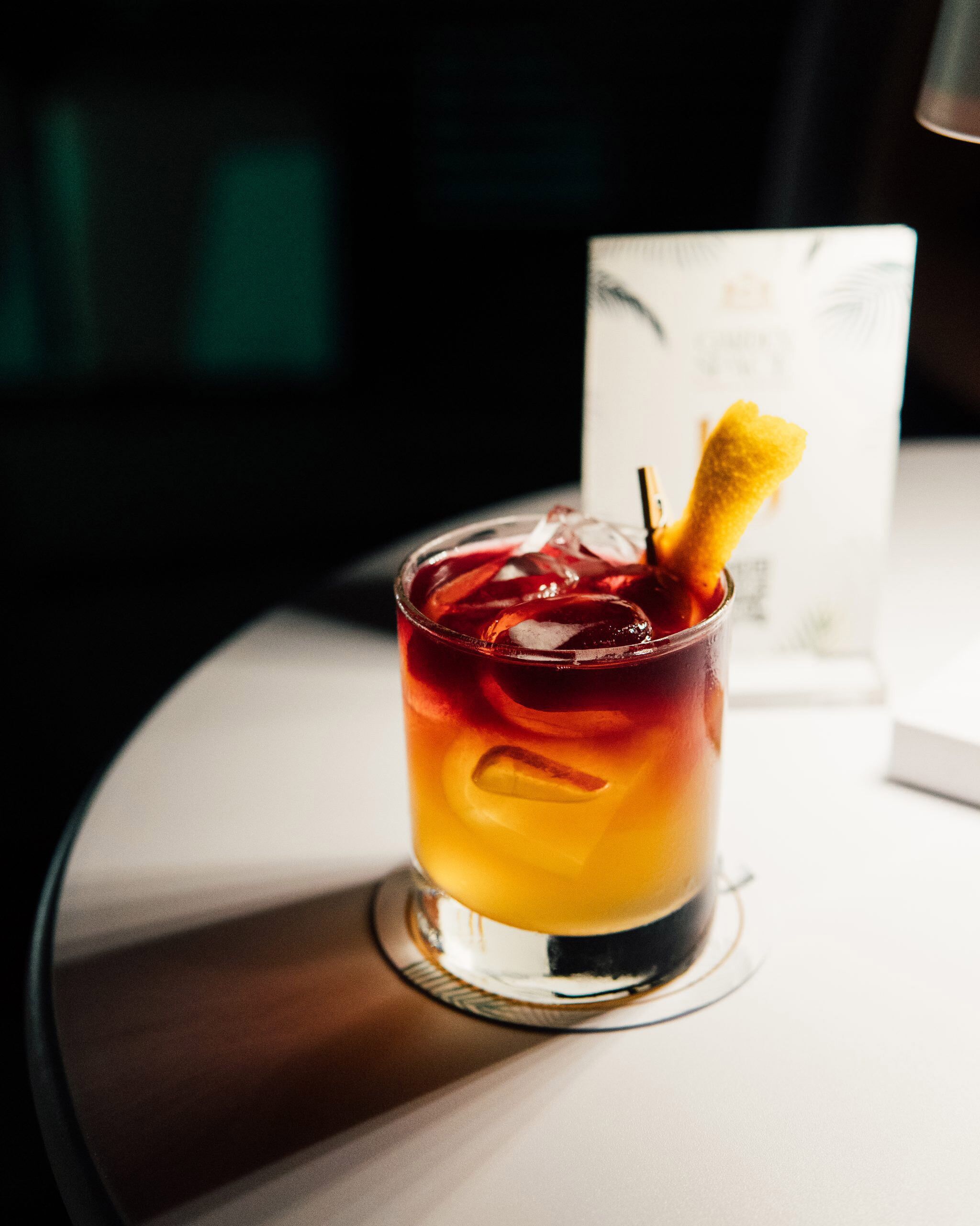 A glass with a layered drink and a lemon slice, placed on a table with a white surface.