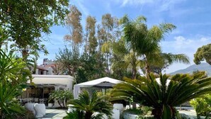 A garden with a covered outdoor seating area, a white building, and palm trees. - Relais Corte degli Aragonesi (Ischia)