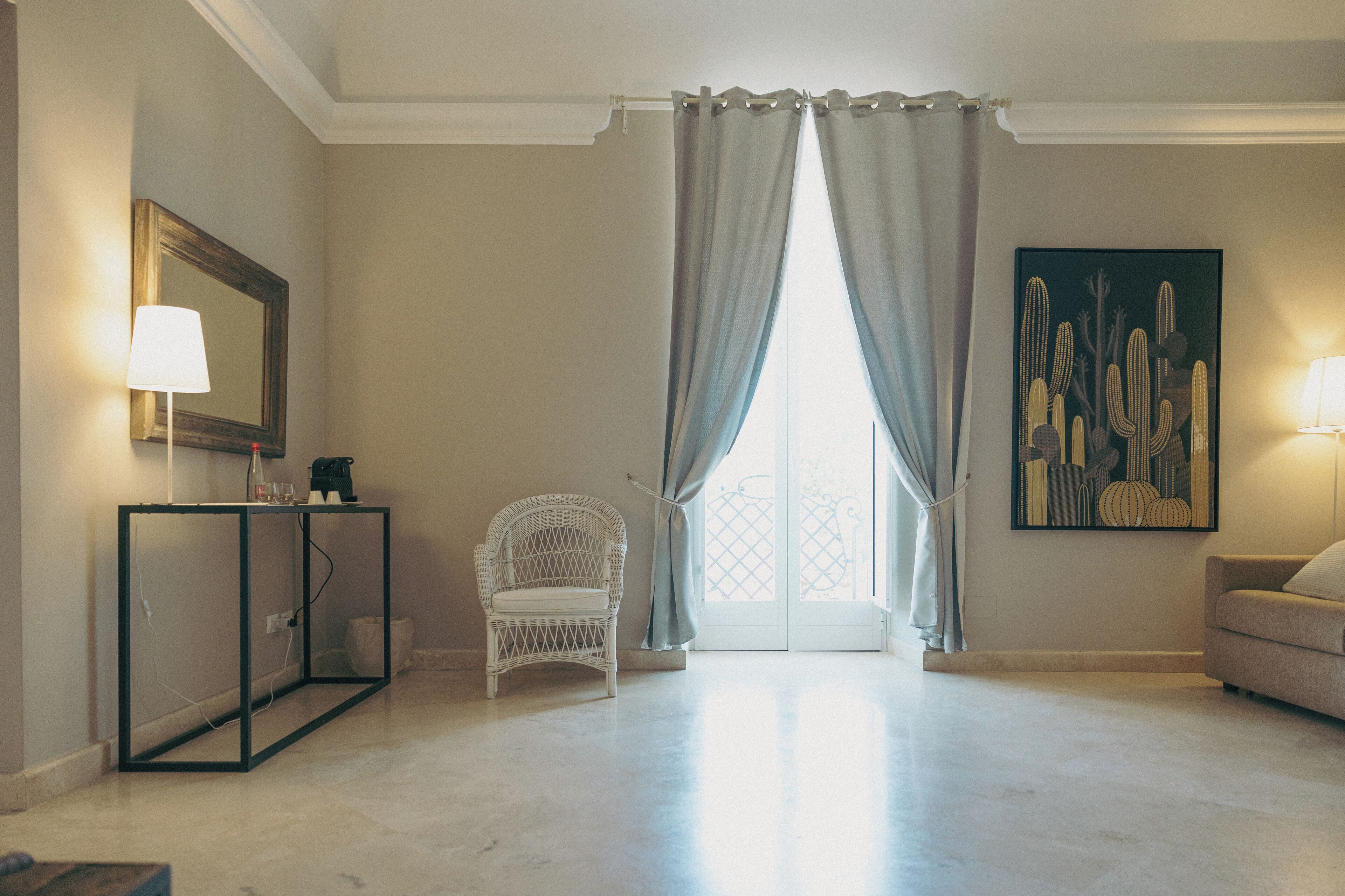 Una sala de estar con una butaca de mimbre blanco, un sofá, una mesa con lámpara, un cuadro y un amplio ventanal con cortinas.
