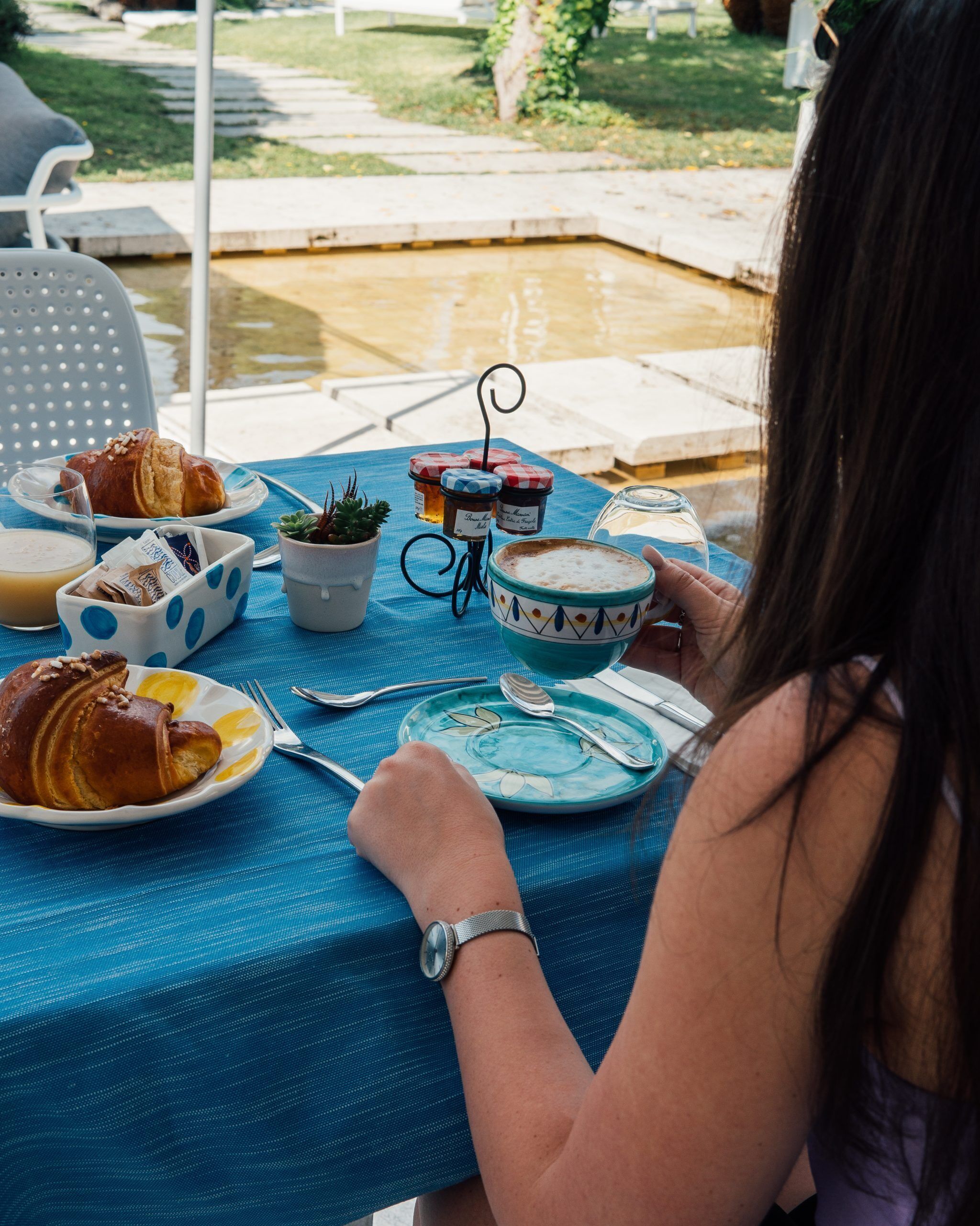A table set with food and drinks, a watch on a wrist, and a garden view in the background.