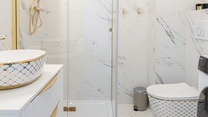 Une salle de bain moderne dotée d’une douche à parois de verre, d’un sol en marbre blanc et d’un lavabo rond avec un receveur à motifs dorés et blancs.