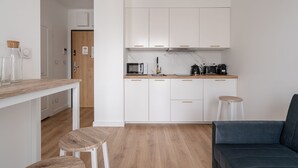 Une cuisine compacte avec des meubles blancs, un plan de travail en bois et une machine à café. On y trouve une table à manger en bois, accompagnée de deux tabourets, ainsi qu’un canapé de couleur foncée.
