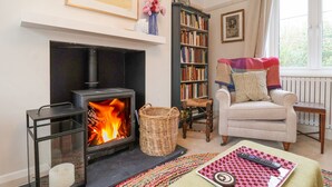 A cozy living room with a fireplace, a bookshelf, a comfortable armchair, and a window with curtains.