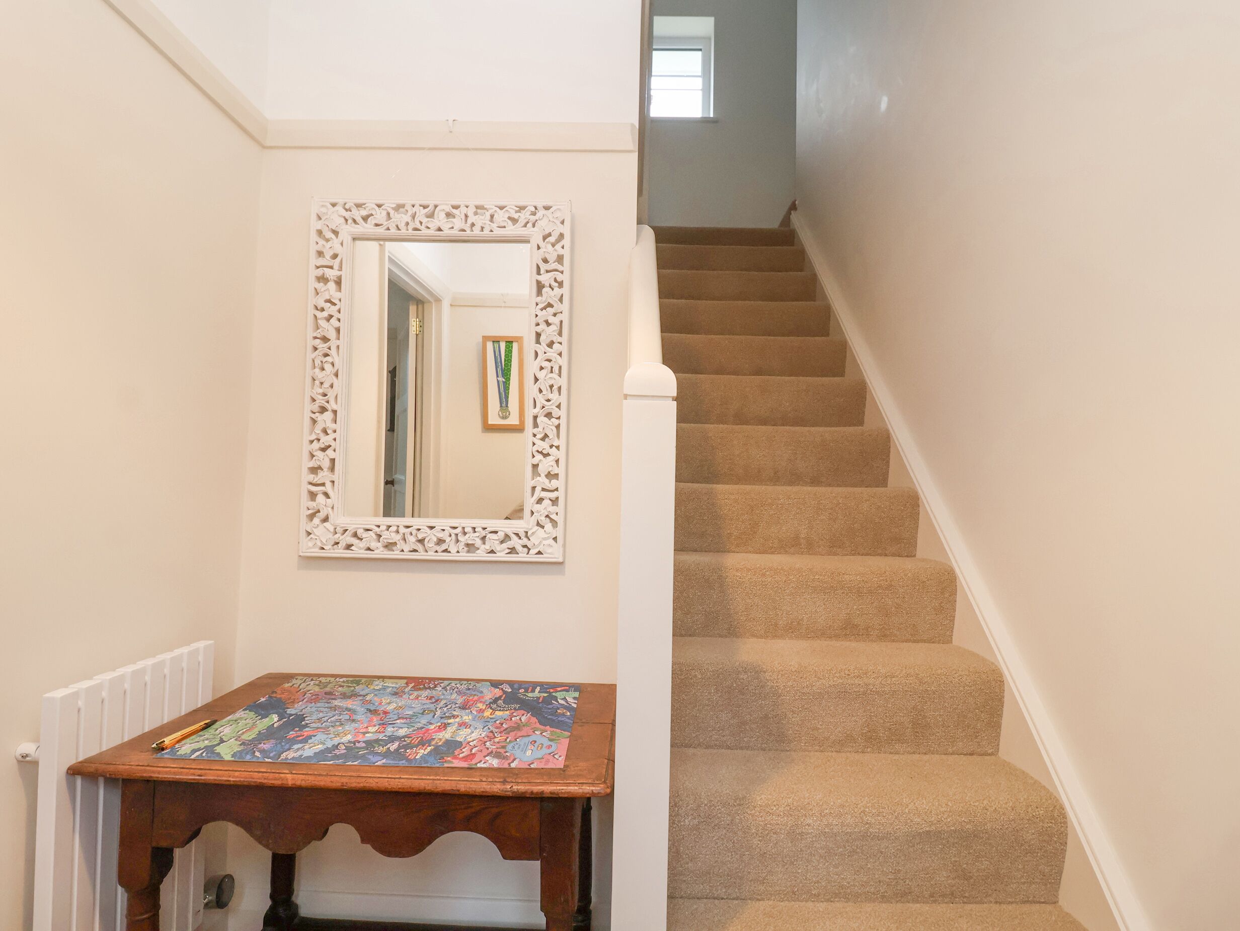 Un couloir avec un escalier recouvert de moquette, une table en bois sur laquelle est posé un jeu de puzzle et un miroir décoratif.