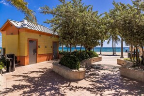 Un bâtiment à la façade jaune, avec une porte et une petite fenêtre. On y voit des arbres et une zone pavée avec un banc.
