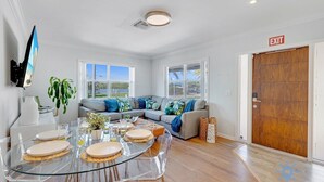 A modern living room with a glass dining table, grey sectional sofa, and a wooden door marked "EXIT".